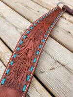 A close-up of a brown leather strap with intricate patterns and teal accents lying on a wooden surface. The strap has detailed carvings and stitching along the edges, showcasing craftsmanship and artistry.