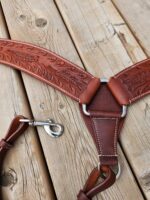 A detailed leather horse harness with intricate engravings and metal rings is laid out on a wooden surface. The harness shows fine craftsmanship with stitching and a clasp attachment.