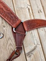 Close-up of a leather harness with intricate embossing on a wooden surface. The harness features detailed patterns, sturdy stitching, and metal rings connecting its parts. The wood background has visible grain and weathering, adding a rustic feel to the scene.