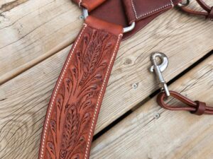 Close-up photo of a handcrafted brown leather horse breast collar with intricate floral tooling displayed on a wooden surface. It includes a metal ring and a snap hook for securing. The detailed stitching enhances the quality and craftsmanship of the piece.