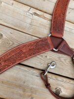 A close-up shot of a leather horse harness with intricate floral engravings on a wooden surface. The harness features reinforced stitching and a metallic clasp on the strap, highlighting its sturdy craftsmanship.