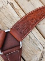 A close-up photograph of a richly textured leather belt with intricate leaf-like engravings. The belt is partially looped through a metal ring, lying on a weathered wooden surface. The craftsmanship details with fine stitching are prominently visible.