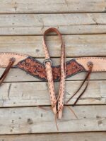 A detailed leather breast collar with intricate floral tooling and black accent designs is laid out on a wooden surface. The collar has adjustable straps and visible stitching, showcasing craftsmanship. It’s designed for equestrian use.