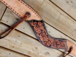 A detailed, handcrafted leather belt with intricate floral patterns rests on a rustic, wooden surface. The belt features a combination of light and dark brown leather with decorative stitching and metal studs. A leather strap is threaded through a hole at one end.