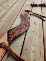 Close-up of a tooled leather belt with intricate floral patterns, laid out on a wooden surface. The belt features shades of brown with detailed carving and lacing, and has leather straps and metal buckles.