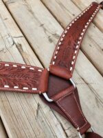 A close-up of a handcrafted leather strap with intricate floral patterns and white stitching. The strap features silver metal rings connecting its sections. The background consists of wooden planks with visible grain and wear.