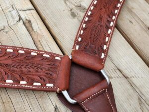 A close-up of a handcrafted leather strap with intricate floral patterns and white stitching. The strap features silver metal rings connecting its sections. The background consists of wooden planks with visible grain and wear.