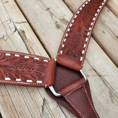 A close-up of a handcrafted leather strap with intricate floral patterns and white stitching. The strap features silver metal rings connecting its sections. The background consists of wooden planks with visible grain and wear.
