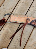 Close-up of a brown leather belt with intricate stitching and decorative elements placed on a wooden surface. The belt features turquoise stitching along the edges and a silver ring with a brown leather strap threaded through it.