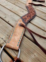 A detailed photo of a leather horse saddle stirrup strap with intricate floral tooling. The strap features blue stitching and metal hardware, laying on a wooden plank surface. Brown leather laces are visible, adding to the design's rustic appearance.