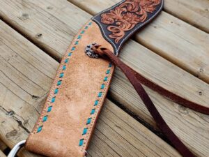 A detailed photo of a leather horse saddle stirrup strap with intricate floral tooling. The strap features blue stitching and metal hardware, laying on a wooden plank surface. Brown leather laces are visible, adding to the design's rustic appearance.