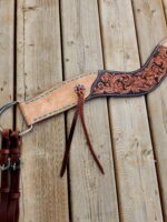 A brown leather horse cinch with intricate floral tooling and turquoise blue stitching lies on a wooden surface. The cinch has leather straps and metal hardware attached to it.