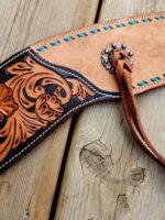 A close-up of a decorative leather belt with intricate floral embossing and turquoise stitching. The belt features a metal concho with a leather strap looped through it, all resting on a wooden surface.