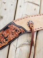 A close-up of a handmade leather belt with intricate floral tooled patterns. The belt features a decorative silver concho and leather lace detailing. It is displayed against a rustic wooden background.