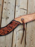 A close-up of a handmade leather belt with intricate floral tooling in black and brown. The belt lies on a wooden surface. It features a decorative metal concho with laces threaded through it, and stitching along the edges.