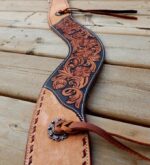 An intricately designed leather belt with a floral motif carved into a dark brown panel is displayed on a wooden surface. The belt has a decorative metal concho and leather ties, showcasing detailed craftsmanship.