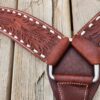Close-up of a brown leather strap with intricate embossed floral designs, white stitching, and metal hardware. The strap is laid on a wooden surface with weathered planks, showing the texture and detail of both materials.