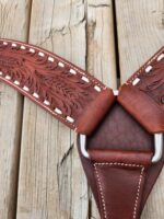 Close-up of a brown leather strap with intricate embossed floral designs, white stitching, and metal hardware. The strap is laid on a wooden surface with weathered planks, showing the texture and detail of both materials.