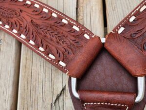 Close-up of a brown leather strap with intricate embossed floral designs, white stitching, and metal hardware. The strap is laid on a wooden surface with weathered planks, showing the texture and detail of both materials.