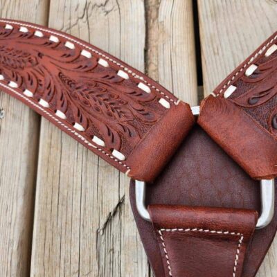 Close-up of a brown leather strap with intricate embossed floral designs, white stitching, and metal hardware. The strap is laid on a wooden surface with weathered planks, showing the texture and detail of both materials.