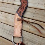 A close-up image of a leather piece featuring intricate floral tooling along the top edge and detailed stitching along the sides. A metal ring and leather strap are attached at the bottom. The item is laid out on a wooden surface.