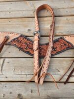 An intricately designed leather rifle sling with detailed floral carvings and a silver buckle, placed on a wooden surface. The sling has adjustable straps with leather ties at the ends.