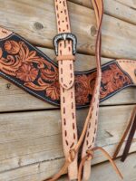 A close-up of a rustic leather guitar strap with intricate carved floral designs. The strap has a mix of natural tan and dark brown hues, with red stitching and a detailed, silver-toned buckle. The background is a wooden surface.