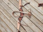 A tan and black leather horse breast collar with intricate tooling and blue stitching lies on a wooden surface. The collar has multiple straps and metal rings for attaching to a saddle, showcasing detailed craftsmanship.