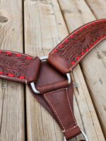 A close-up of a detailed leather horse tack piece lying on a wooden surface. The leather is dark brown with intricate floral engraving and red stitching around the edges. Metal rings and white stitching are also visible.