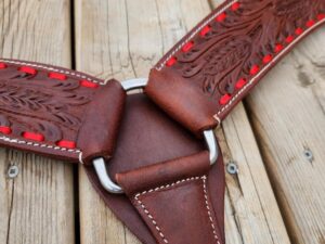 A close-up of a detailed leather horse tack piece lying on a wooden surface. The leather is dark brown with intricate floral engraving and red stitching around the edges. Metal rings and white stitching are also visible.