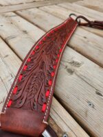 A close-up of a decorative leather belt with intricate carvings and red accents. The belt is laid out on a light wooden surface, showcasing its detailed craftsmanship and vibrant coloring.