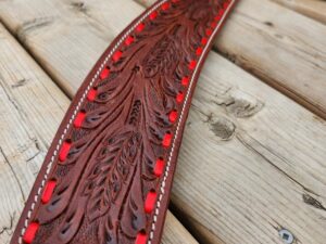 A close-up of a decorative leather belt with intricate carvings and red accents. The belt is laid out on a light wooden surface, showcasing its detailed craftsmanship and vibrant coloring.