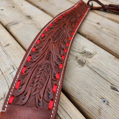 A close-up of a decorative leather belt with intricate carvings and red accents. The belt is laid out on a light wooden surface, showcasing its detailed craftsmanship and vibrant coloring.