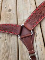 Close-up of intricately tooled leather tack with red stitching and silver rings, laying on a wooden surface. The leather features a detailed leaf pattern and is part of a horse's breastcollar.