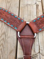 A close-up image of intricately designed leather straps, connected by a metallic ring on a wooden surface. The leather is embossed with a floral pattern and accented with blue inlays, showcasing fine craftsmanship and detailed stitching.