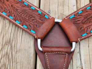 A close-up image of intricately designed leather straps, connected by a metallic ring on a wooden surface. The leather is embossed with a floral pattern and accented with blue inlays, showcasing fine craftsmanship and detailed stitching.