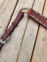 Close-up of a brown leather belt with a decorative silver buckle and a leather sheath attached to a metal ring. The sheath has turquoise accents and intricate tooling patterns. The items are laid on a wooden surface with visible grain and knots.