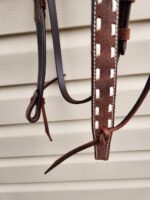 Close-up image of a leather horse bridle with white stitching detail, hanging against a beige ribbed wall. The leather has brown and black straps, with some ends tied into knots or loops. The bridle showcases intricate craftsmanship and sturdy design.