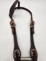 A brown leather bridle piece with intricate silver and copper decorative buckles and accents. The leather is stitched with white thread, and the metal bits are polished and shiny. The photo is taken on a neutral, light background.