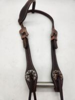 A close-up image of a horse bridle made of dark brown leather. The bridle features intricate silver decorations and detailed stitching along the edges. The metal bit is visible at the bottom of the bridle. The background is a plain, light surface.