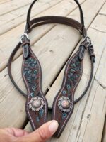 A hand is holding a piece of a brown leather bridle adorned with intricate turquoise embroidery and two metal conchos with a floral design. The bridle is laid out on a wooden surface.