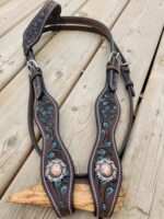 A detailed leather horse bridle featuring intricate turquoise-colored patterns and decorative metal conchos with floral engravings, is laid out on a rustic wooden surface. The bridle includes buckles and straps for adjustment.