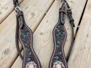 A detailed leather horse bridle featuring intricate turquoise-colored patterns and decorative metal conchos with floral engravings, is laid out on a rustic wooden surface. The bridle includes buckles and straps for adjustment.