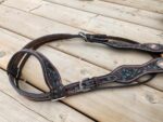 A brown leather horse bridle with turquoise floral engravings is laid out on a wooden deck. The bridle features silver buckles and detailed stitching. The wood planks are light-colored and slightly weathered.