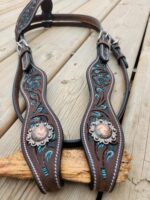 A detailed leather horse bridle with turquoise accents and intricate floral patterns. The bridle is decorated with conchos and white stitching. It rests on a wooden surface.