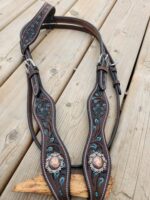 Leather horse tack with ornate turquoise and silver detailing rests on a wooden surface. The leather features intricate carvings and two large decorative conchos. The background is a wooden deck that shows signs of wear.