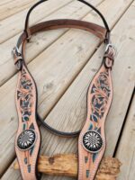 A decorative leather headstall with intricate floral tooling and turquoise inlays is displayed on a wooden surface. The headstall features silver conchos with sunburst designs and adjustable buckles on both sides.