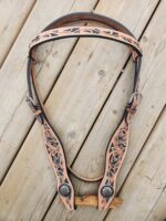 Close-up of an intricately designed leather headstall with floral patterns in black and turquoise. The headstall, likely used for horse tack, is laid out on a wooden surface, showcasing its detailed craftsmanship with silver-tone accents.