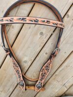 A leather rifle sling with intricate carved patterns, featuring teal, black, and tan colors, lies coiled on a wooden surface. The sling has adjustable straps and a soft, padded section for shoulder comfort.