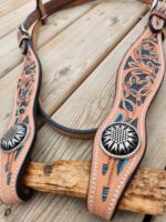 A intricately crafted leather horse bridle is decorated with teal and blue accents, floral designs, and silver conchos. It is resting on a wooden surface.